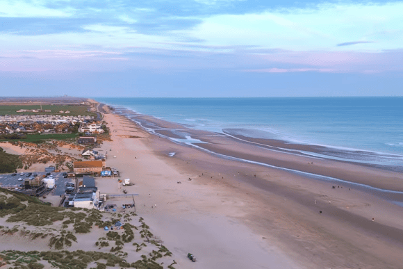 Aerial panorama of Camber Sands with miles of golden sandy beach, rolling sand dunes, tidal sandbars, and the English Channel stretching along the East Sussex coastline at sunset.