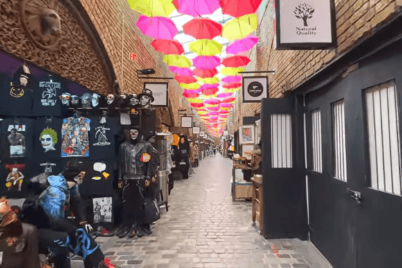 Colourful umbrella-covered walkway inside Camden Stables Market in London, lined with independent stalls selling clothing, art, gifts and accessories.
