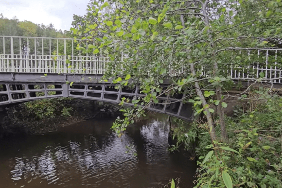 Cast-iron Cantlop Bridge spanning a narrow river, with white railings above and leafy trees overhanging the water.