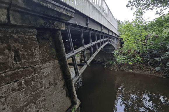 Underside view of Cantlop Bridge showing the cast-iron lattice arch and stone abutment above the River Rea, framed by dense greenery.