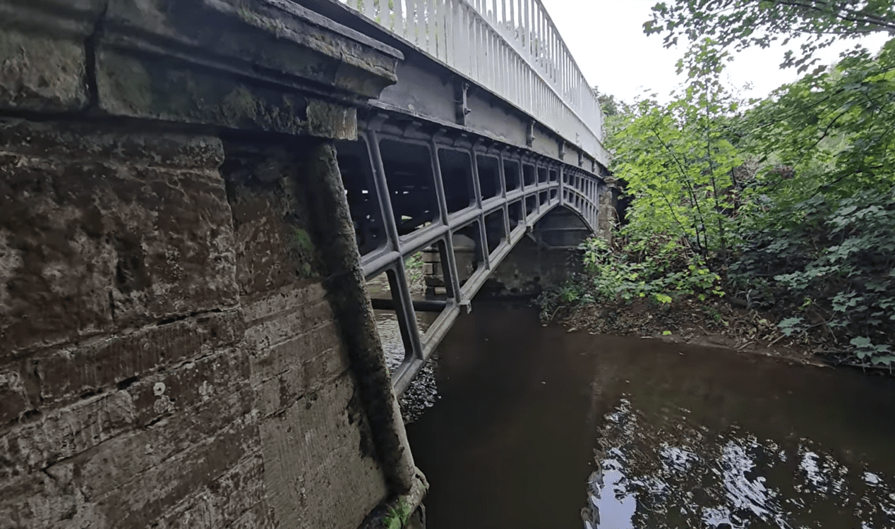 Underside view of Cantlop Bridge showing the cast-iron lattice arch and stone abutment above the River Rea, framed by dense greenery.
