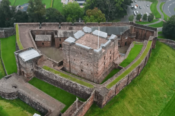 Aerial view of Carlisle Castle showing the central stone keep, curtain walls, bastions, and surrounding green lawns within the city.