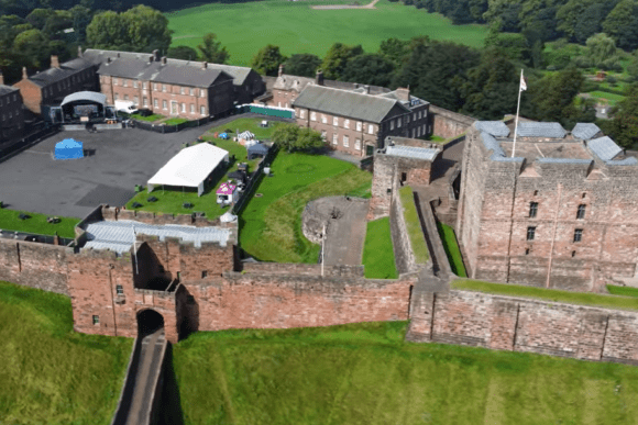 Aerial view of Carlisle Castle showing the stone keep, curtain walls, inner courtyard buildings, and green lawns beyond the fortifications.