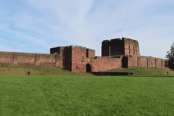 Wide view of Carlisle Castle showing the red sandstone curtain walls, central keep, and grassy outer bailey under a blue sky.