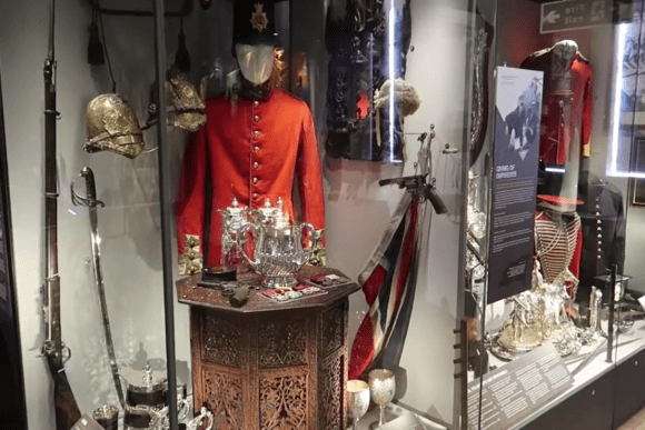 Museum display at Carlisle Castle showing a red military tunic, ceremonial silverware, swords, helmets, and historic uniforms behind glass.