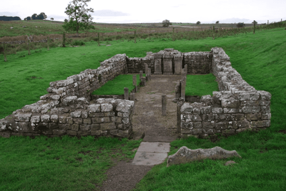 Stone foundations of the Temple of Mithras at Carrawburgh Roman Fort, with low walls and altar bases set in grassy countryside.