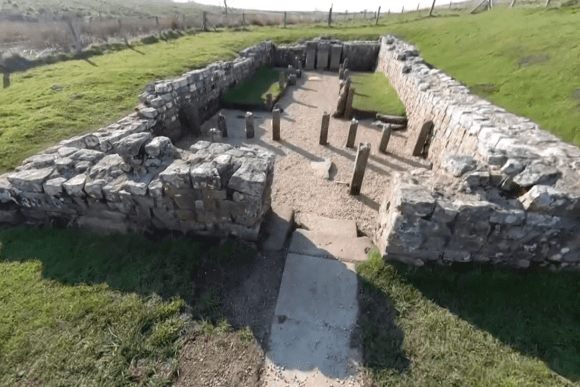 Rectangular stone remains of the Temple of Mithras at Carrawburgh Roman Fort, showing altar bases and low walls set in open grassland.