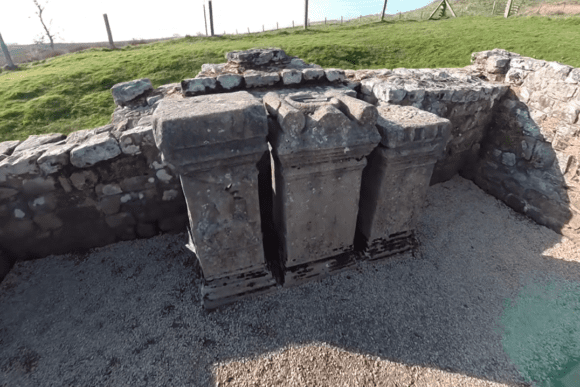 Stone altar bases inside the Temple of Mithras at Carrawburgh Roman Fort, standing against low stone walls on Hadrian’s Wall.
