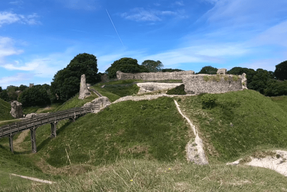 Wide view of Castle Acre Castle showing the grassy motte, stone ruins on the summit, and the wooden footbridge leading to the Bailey Gate under a blue sky.