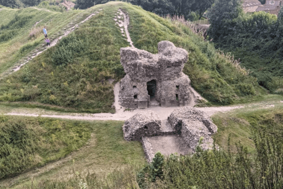 Stone Bailey Gate ruins at Castle Acre Castle set into a grassy earthwork, with footpaths climbing the surrounding motte and visitors walking along the ridge.