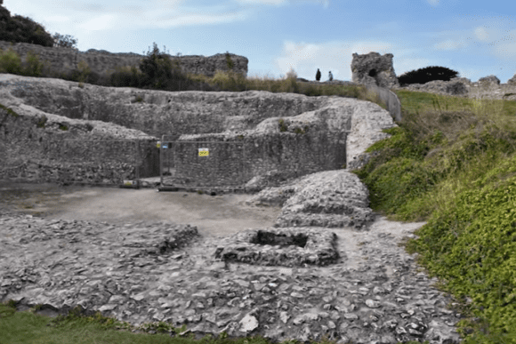 Excavated stone foundations and earthworks inside Castle Acre Castle, with low masonry walls, sloping banks, and ruined sections visible on the hilltop beyond.