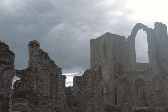 Ruined stone walls and arches of Castle Acre Priory silhouetted against a dramatic, overcast sky.