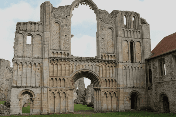 The monumental west front of Castle Acre Priory, showing tall Norman stone arches, layered arcading, and weathered ruins set on a grassy lawn.