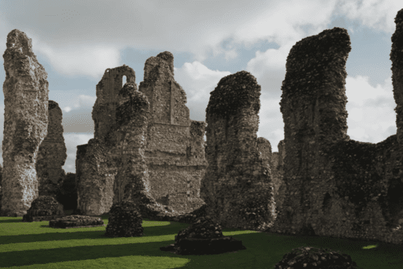 Tall, weathered flint-and-stone columns and walls of Castle Acre Priory rising from a grassy cloister beneath a cloudy sky.