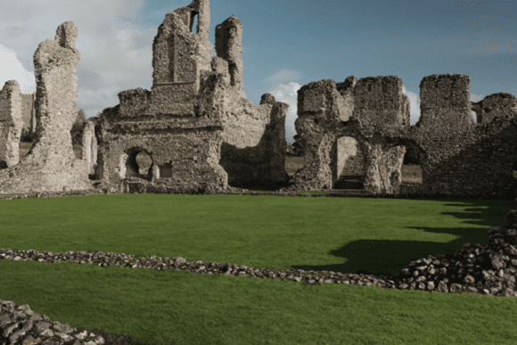 Ruined stone walls and arched openings of Castle Acre Priory surrounding a neatly kept grassy cloister under a blue sky.