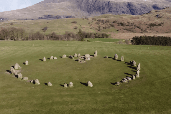 Castlerigg Stone Circle set in an open grassy field, with ancient standing stones arranged in a ring against the dramatic mountain backdrop of the Lake District.