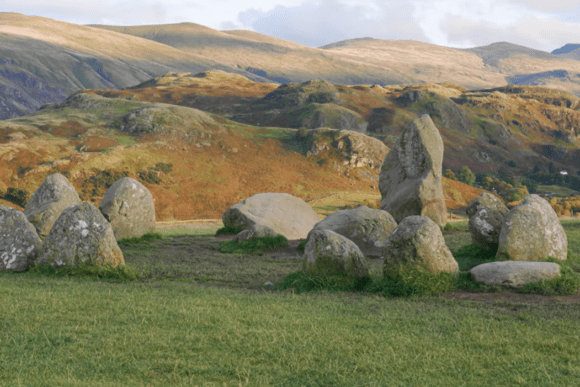 Standing stones of Castlerigg Stone Circle in the foreground, with rolling Lake District fells and autumn-coloured hills stretching into the distance.
