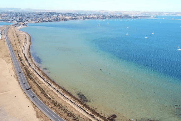 Aerial view of Chesil Beach at the Portland end, showing the sweeping shingle coastline, turquoise waters of Portland Harbour, anchored sailboats, and the Jurassic Coast in Dorset.