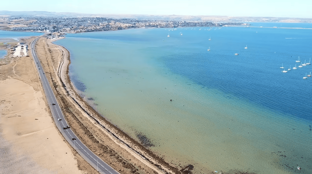 Aerial view of Chesil Beach at the Portland end, showing the sweeping shingle coastline, turquoise waters of Portland Harbour, anchored sailboats, and the Jurassic Coast in Dorset.