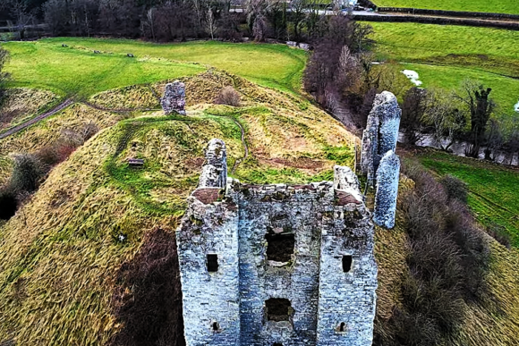 Aerial view of the ruined stone keep of Clun Castle on a grassy hill, with surrounding countryside and winding paths below.