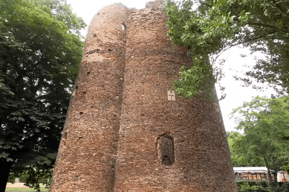 Exterior view of Cow Tower in Norwich, showing the tall circular brick structure surrounded by trees.