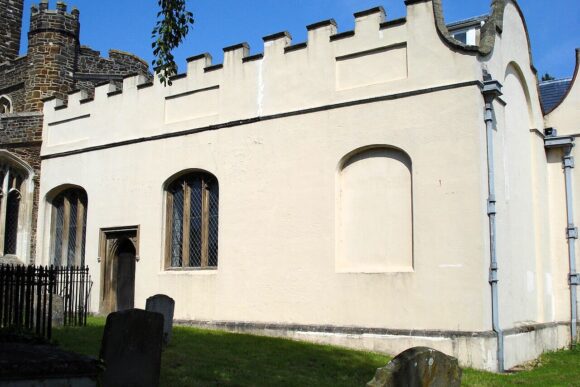 Exterior of the De Grey Mausoleum, a pale stone building with crenellated roofline beside a churchyard.