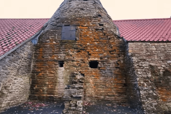 Stone cone and wall of Derwentcote Steel Furnace with small openings and red-tiled roofs on either side.