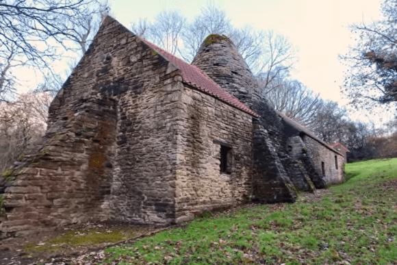 Stone-built Derwentcote Steel Furnace with conical chimney and pitched roof, set on a grassy slope among leafless trees.