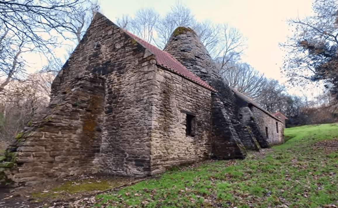Stone-built Derwentcote Steel Furnace with conical chimney and pitched roof, set on a grassy slope among leafless trees.