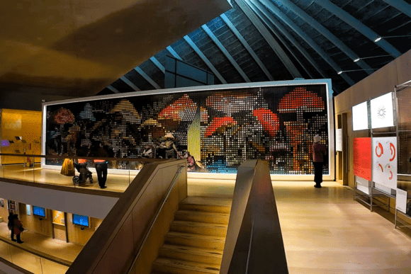 Modern exhibition hall inside the Design Museum in London featuring a large illuminated digital artwork inspired by colourful mushrooms, contemporary gallery architecture, and visitors exploring interactive design displays.