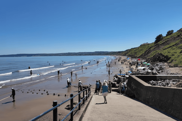 Visitors enjoying Filey Beach in North Yorkshire, with families paddling in shallow waters, sandy shoreline, and scenic coastal cliffs under a bright blue summer sky.