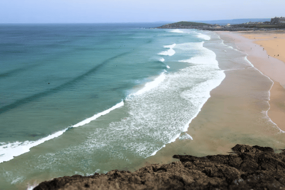 Aerial view of Fistral Beach in Newquay, Cornwall, with rolling Atlantic waves, golden sand, turquoise water, and dramatic rocky cliffs.