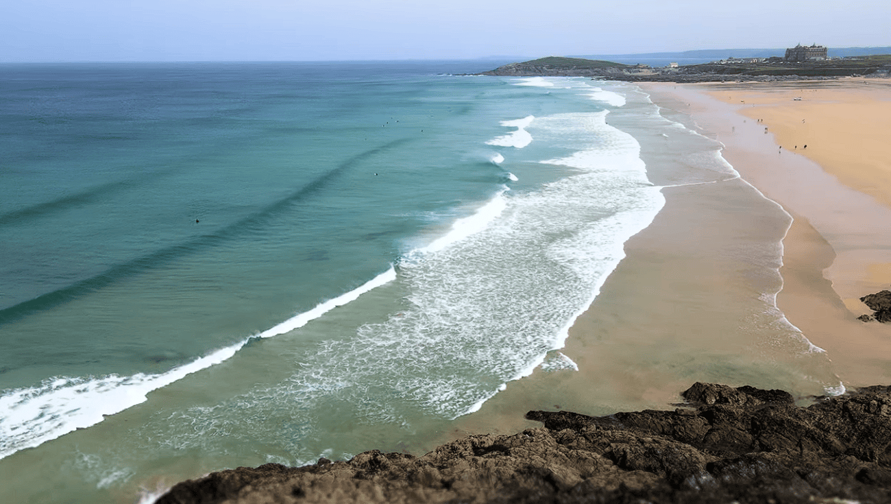 Aerial view of Fistral Beach in Newquay, Cornwall, with rolling Atlantic waves, golden sand, turquoise water, and dramatic rocky cliffs.