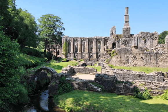 Ruins of Fountains Abbey framed by greenery, with a small stone bridge over a stream in the foreground and arched abbey walls in the background.