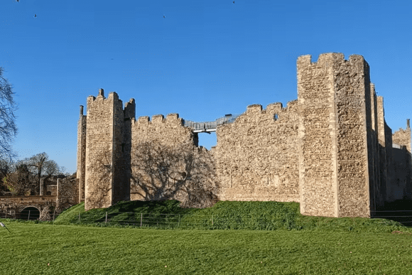 Wide view of Framlingham Castle’s stone curtain walls and towers rising above a grassy field under a clear blue sky.