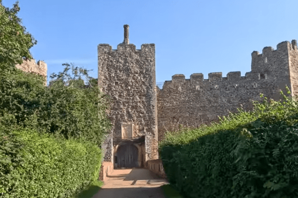 Stone gate tower and curtain wall of Framlingham Castle approached by a narrow path lined with greenery.
