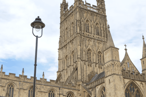 View of Gloucester Cathedral’s ornate stone tower and Gothic architecture rising above surrounding buildings under a cloudy sky.