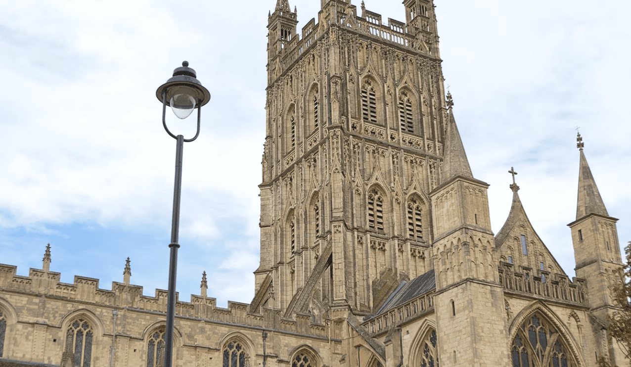 View of Gloucester Cathedral’s ornate stone tower and Gothic architecture rising above surrounding buildings under a cloudy sky.