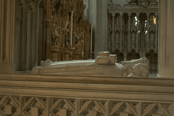 Stone tomb with an effigy inside Gloucester Cathedral, set before an ornate altar and carved choir screen.