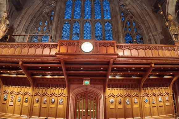 Ornate wooden gallery inside Guildhall London with carved paneling, central clock, stained-glass window, and decorative statues.