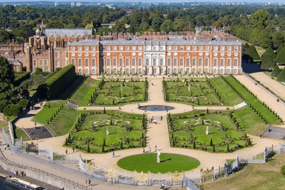 Aerial view of Hampton Court Palace with formal gardens, symmetrical lawns, fountains, and the historic red-brick palace.