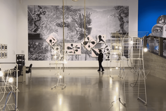 Contemporary mixed-media installation at Hayward Gallery in London featuring illuminated wire-frame sculptures, large-scale monochrome wall art, geometric graphic elements, and visitors exploring the exhibition space.