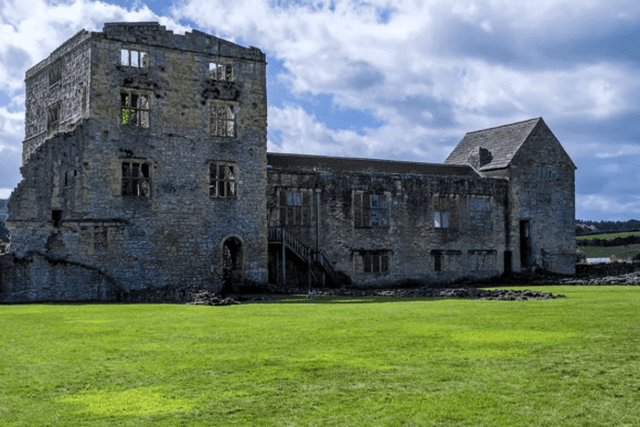 Ruined stone buildings and tower of Helmsley Castle standing beside a grassy courtyard under a cloudy sky.
