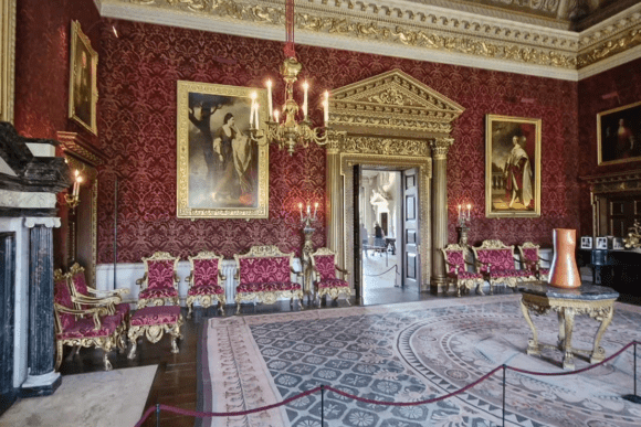 Ornate state room in Houghton Hall with red patterned walls, chandeliers, paintings, and antique chairs.