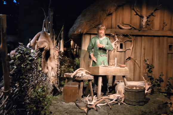 Recreated Viking craftsman workshop at JORVIK Viking Centre in York featuring a life-sized Norse figure carving antlers outside a timber longhouse with authentic Viking tools and animal hides.