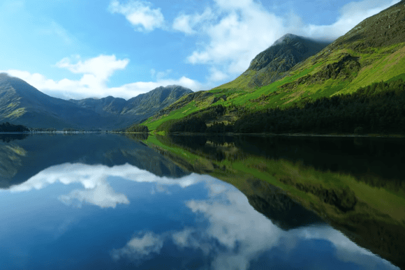 Panoramic view of a tranquil lake in the Lake District National Park reflecting green mountains, forests, and blue skies with scattered clouds on a calm day.