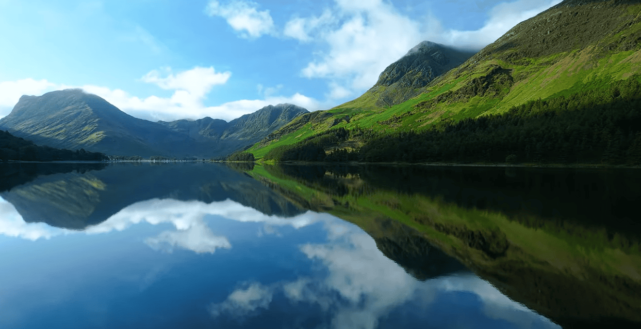 Panoramic view of a tranquil lake in the Lake District National Park reflecting green mountains, forests, and blue skies with scattered clouds on a calm day.