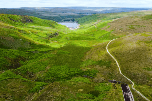 Panoramic view of rolling green hills and winding walking trails in the Lake District National Park leading toward a peaceful reservoir surrounded by dramatic countryside.