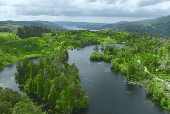 Aerial view of tranquil lakes, lush green forests, and rolling hills in the Lake District National Park, with winding walking trails and distant mountain scenery under dramatic clouds.