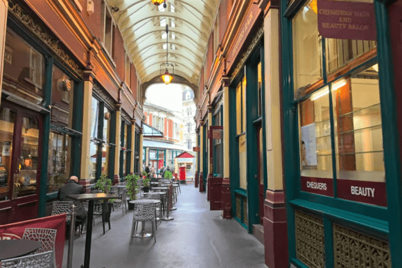 Interior walkway of Leadenhall Market with covered glass roof, shopfronts on both sides, and outdoor café seating.